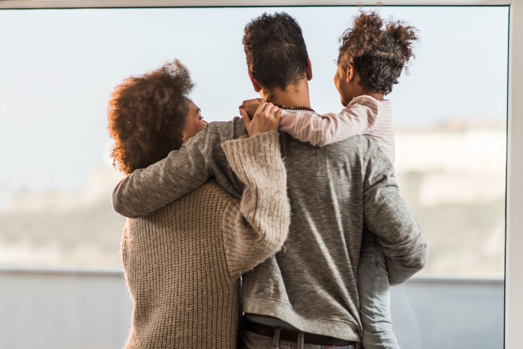 Rear view of embraced African American family looking through the window.