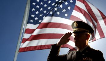 Silhouette of veteran US Army Colonel Chaplain wearing hat and saluting with an American flag flying behind him.