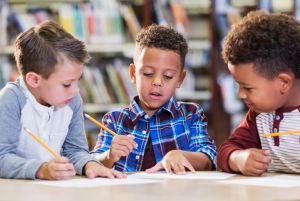 Three multi-ethnic boys in library writing at table