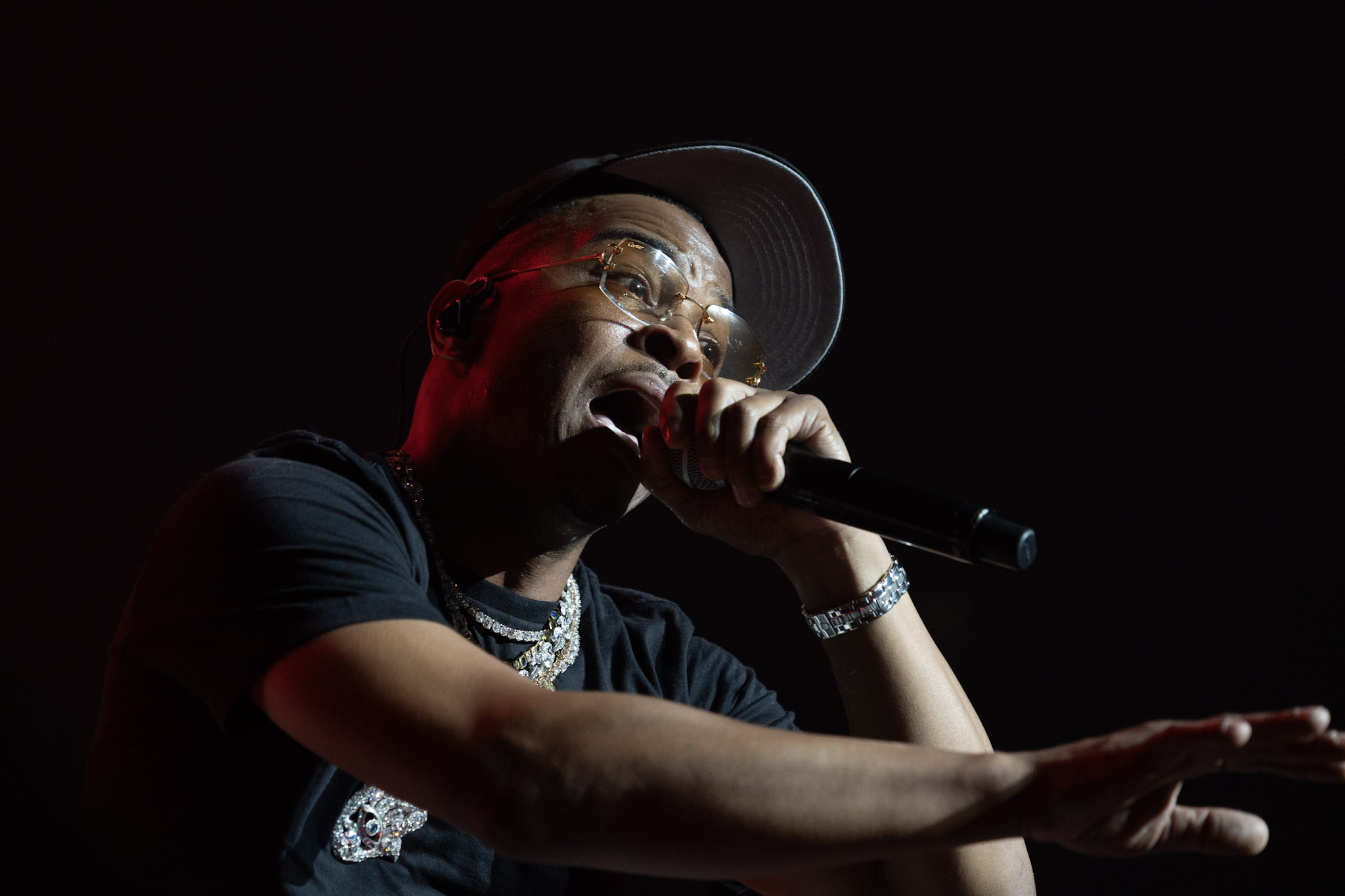 A performer wearing a red hat and sunglasses singing into a microphone on a dark stage.
