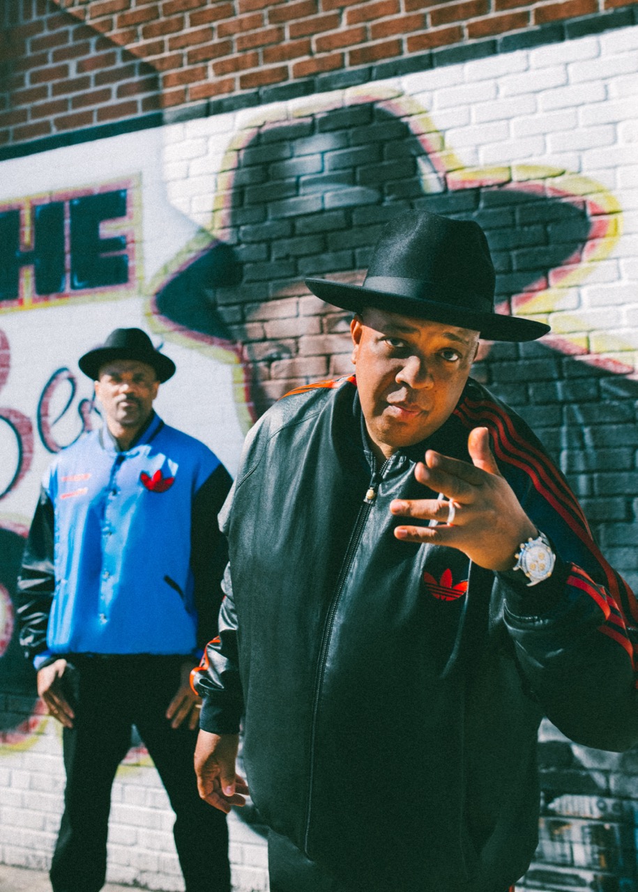 Two men wearing hats and jackets standing in front of a brick wall with graffiti.