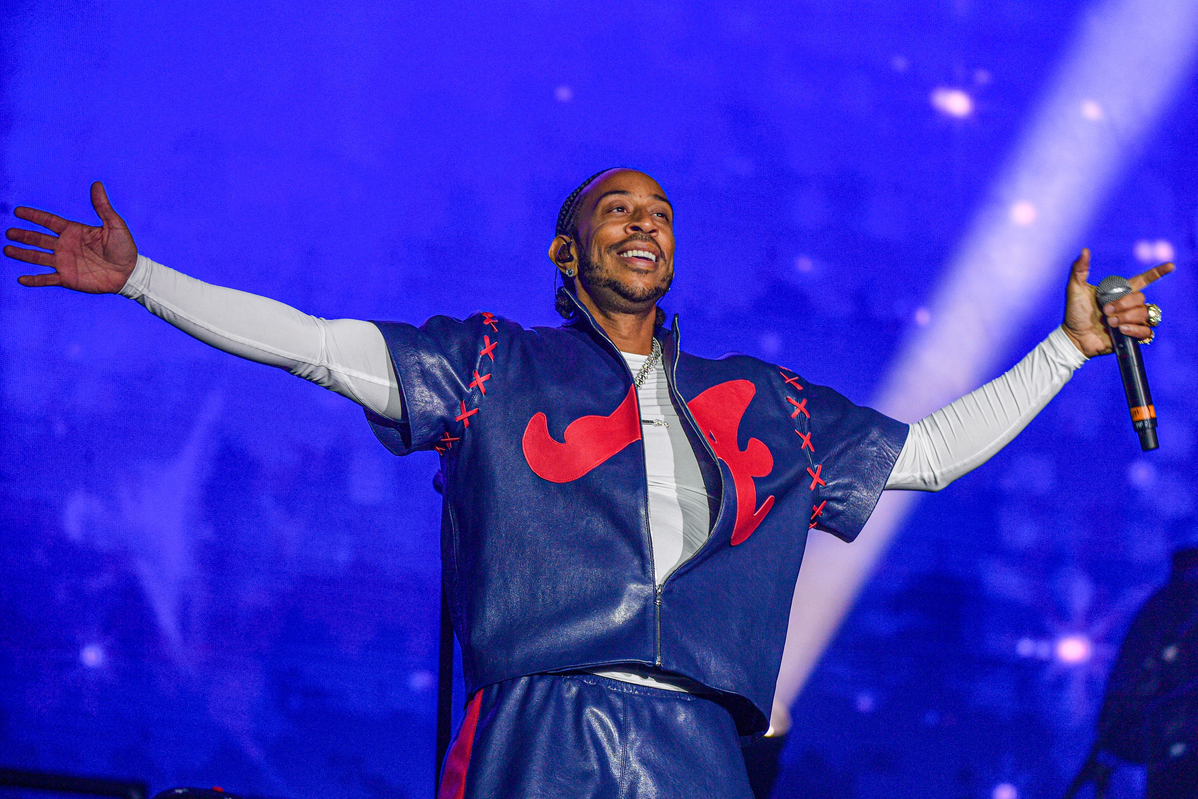 A performer in a blue and red leather outfit with arms raised, performing on a stage against a blue background with lights.