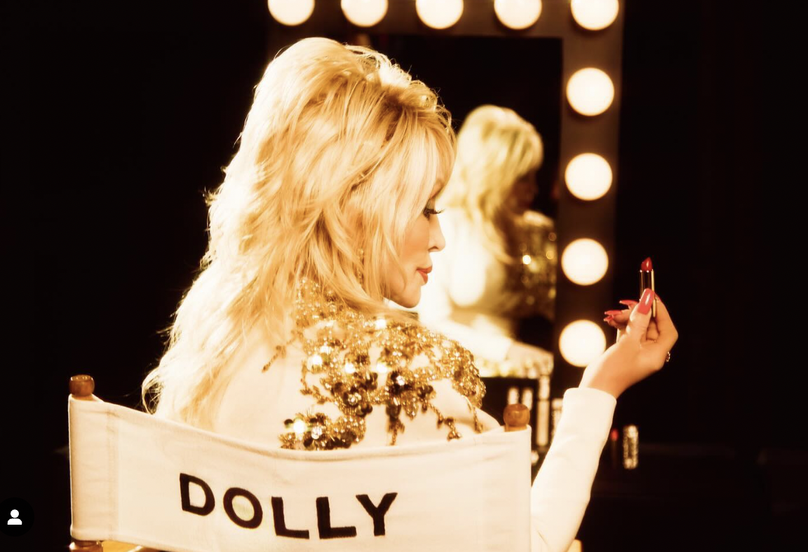 A woman with long blonde hair applying lipstick in front of a lighted mirror, wearing a gold sequined top.