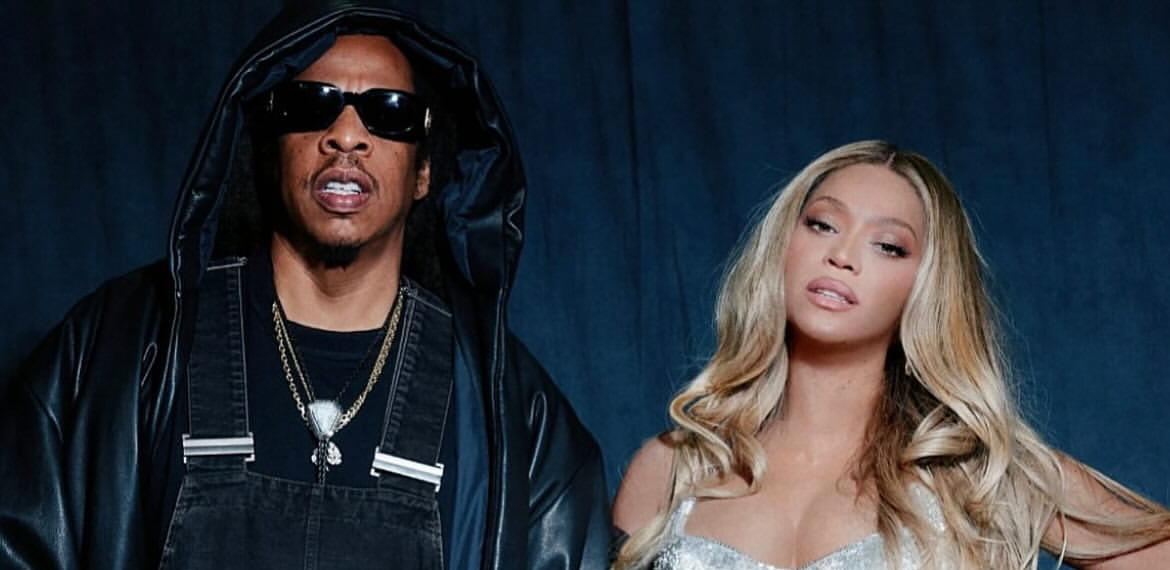 Two people, a man in a black hooded jacket and a woman in a silver sequined outfit, standing together against a dark background.