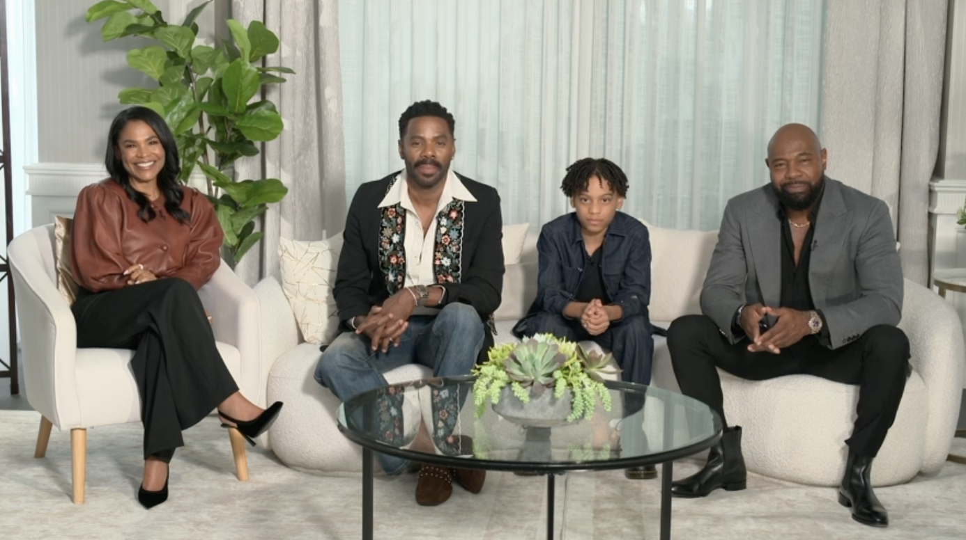 A group of four people, two men and two women, sitting on a couch in a living room setting. The room has a large window and a potted plant on the table in front of them.