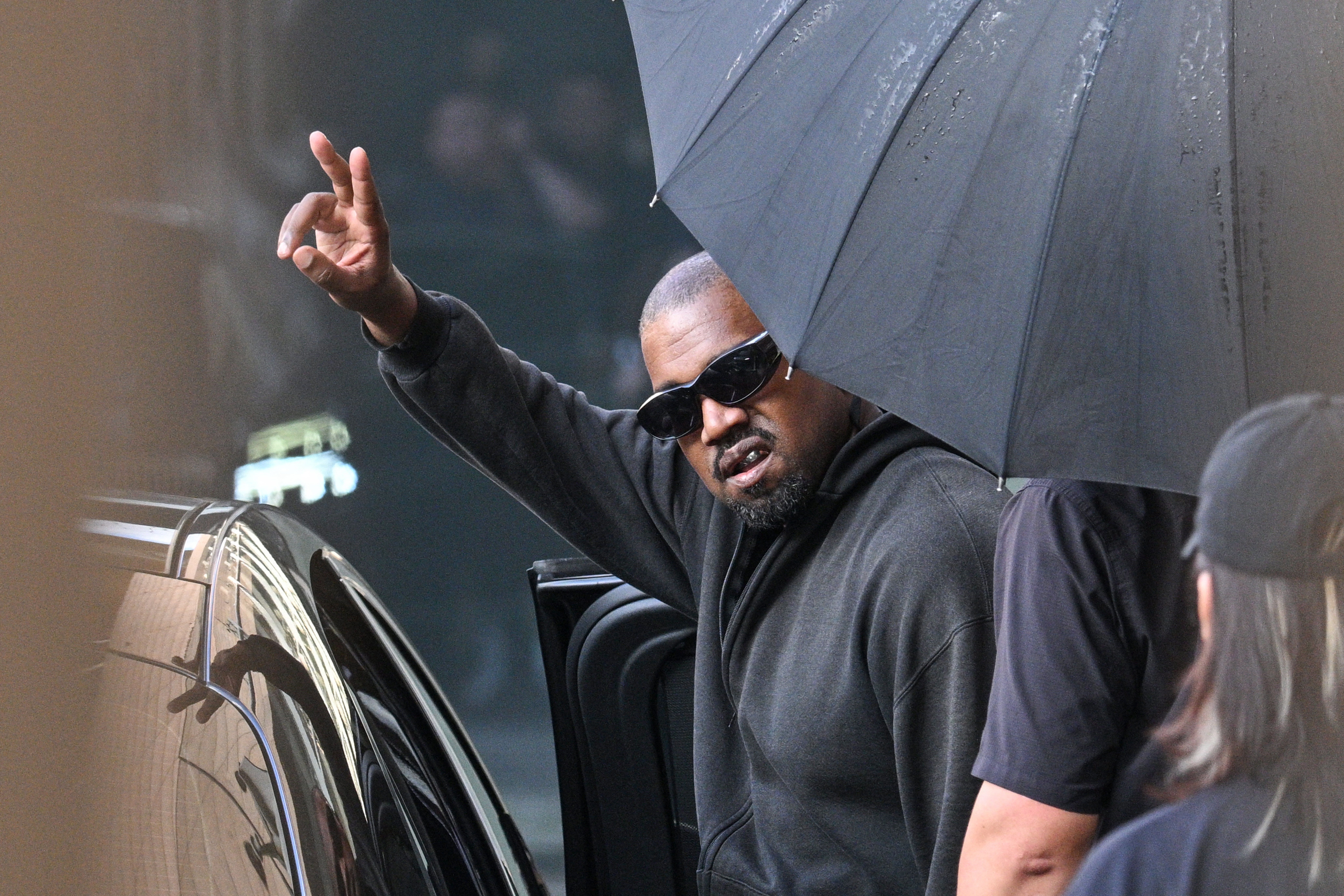 A person wearing sunglasses and a black hoodie gesturing with their hand while holding an umbrella in front of a car.