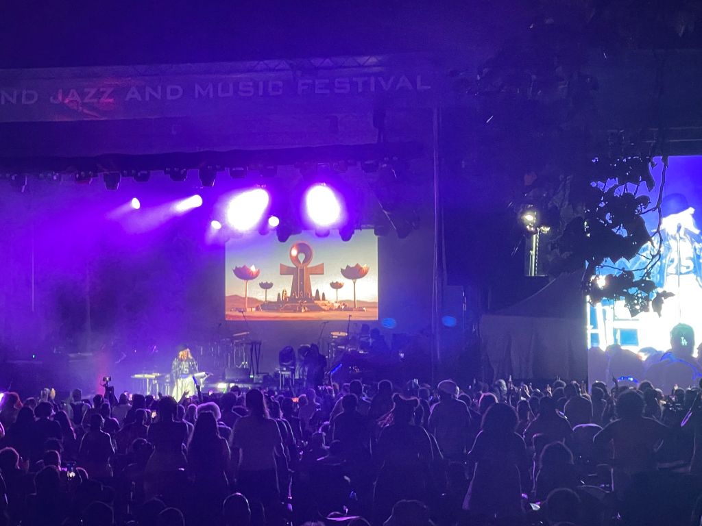Large crowd at a music festival on a stage with colorful lighting and a large screen displaying a logo and text "GRAND JAZZ AND MUSIC FESTIVAL".